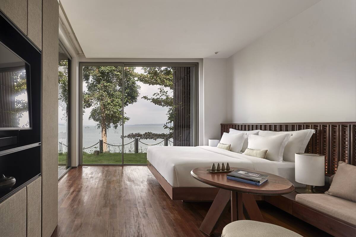 Une chambre d’hôtel moderne avec un grand lit, un canapé, une petite table et un téléviseur. La chambre offre une vue sur la mer et sur un arbre.