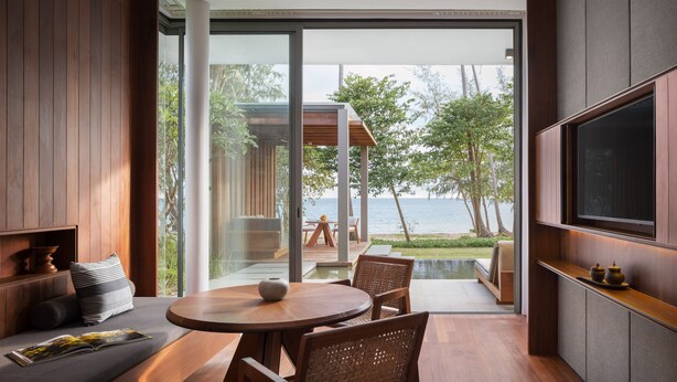Un salon moderne avec une table ronde en bois et des chaises en osier, un canapé avec des coussins, un téléviseur à écran plat fixé au mur et une grande porte vitrée donnant sur une terrasse avec vue sur la mer.