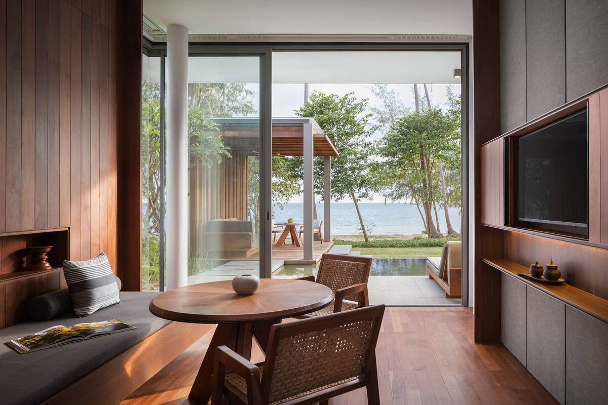 Un salon moderne avec une table ronde en bois et des chaises en osier, un canapé avec des coussins, un téléviseur à écran plat fixé au mur et une grande porte vitrée donnant sur une terrasse avec vue sur la mer.