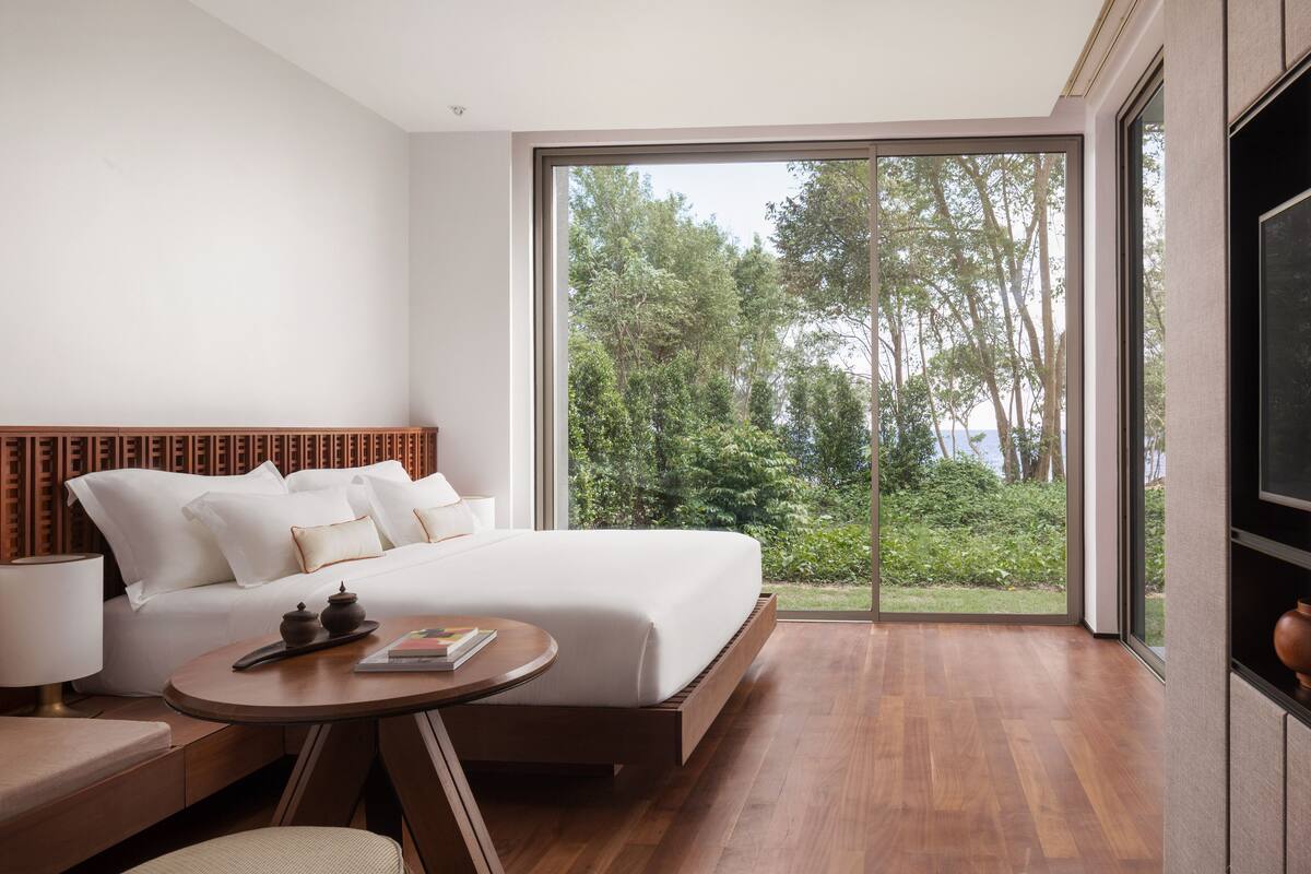 Une chambre à coucher moderne avec un grand lit, une tête de lit en bois, une table ronde en bois et une vue sur un jardin luxuriant, visible à travers une porte vitrée.