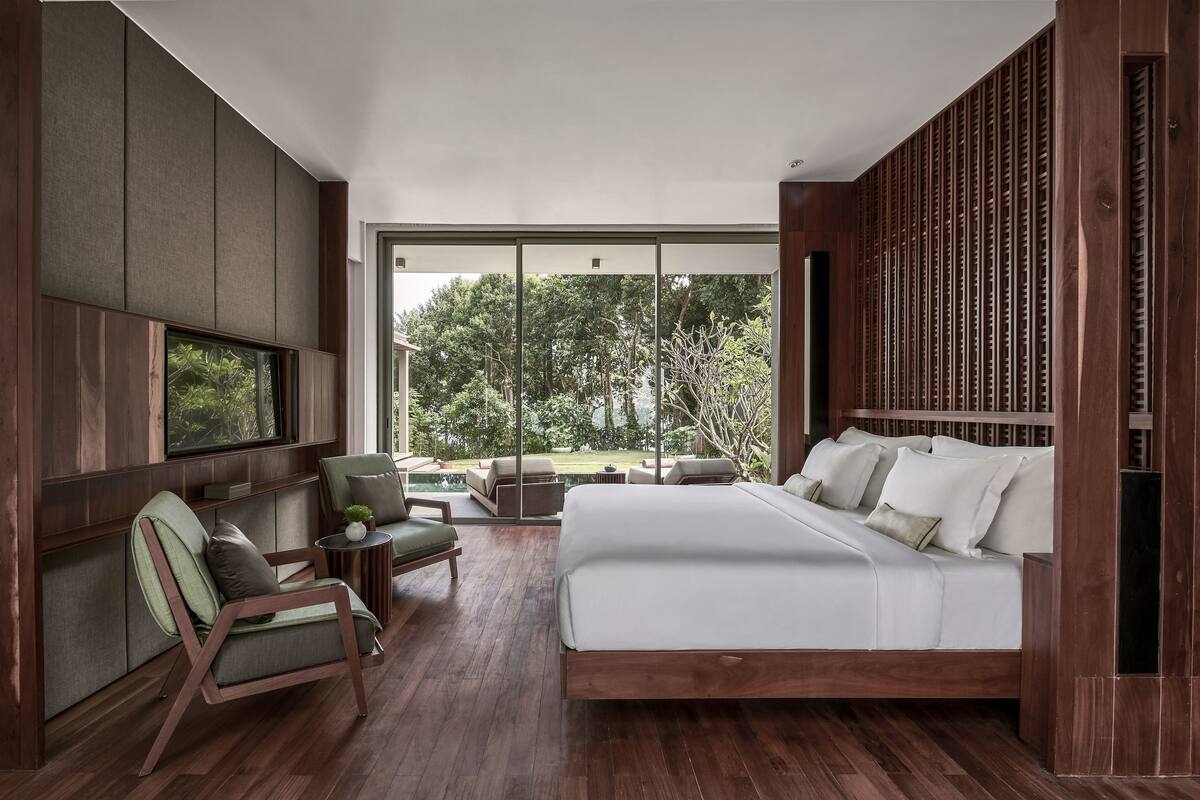 Une chambre moderne dotée d’un grand lit, d’une télévision, d’un coin salon avec des fauteuils et offrant une vue sur la verdure environnante.