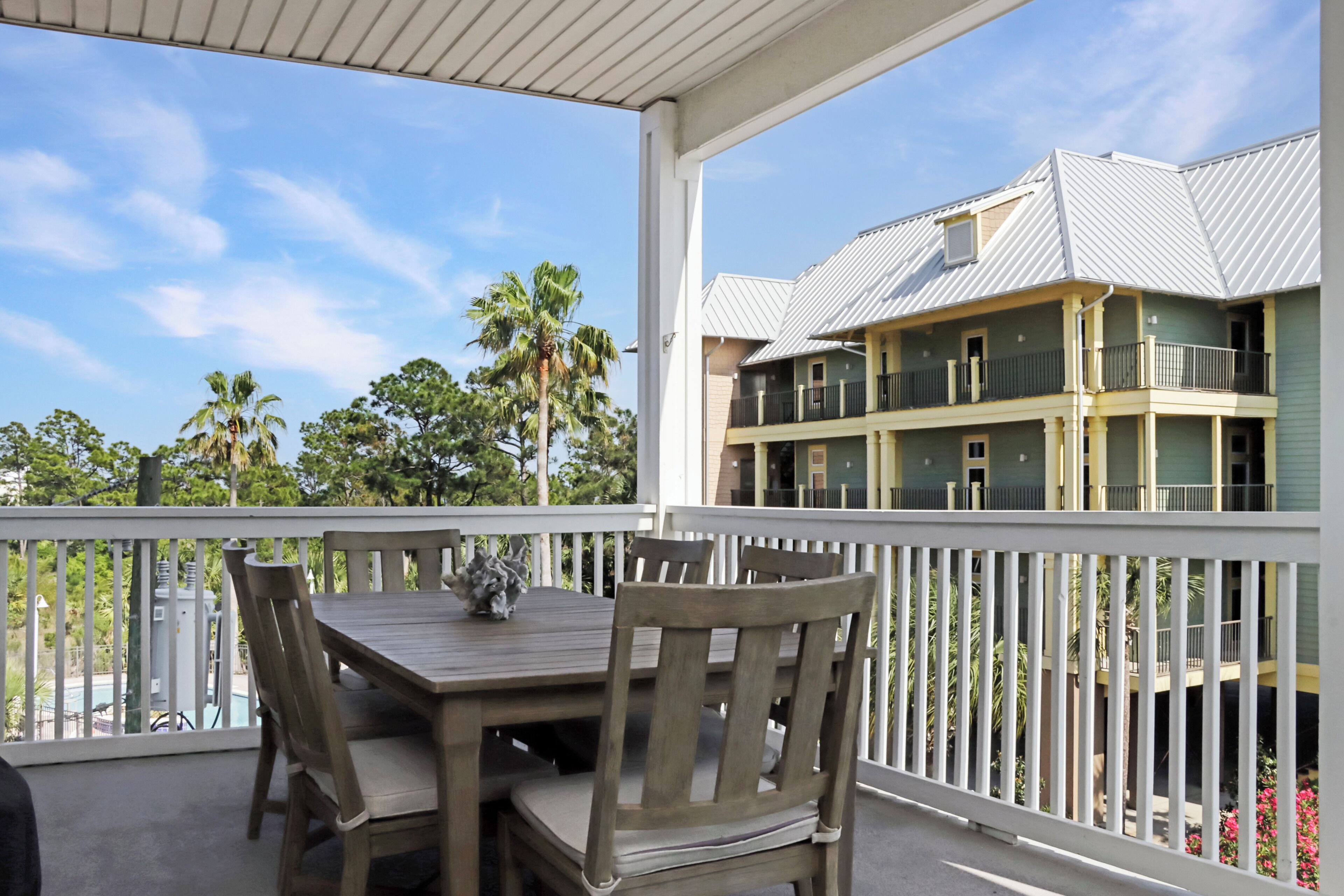 A covered outdoor area with a wooden table and chairs, a white railing, and a view of a multi-story building with balconies.