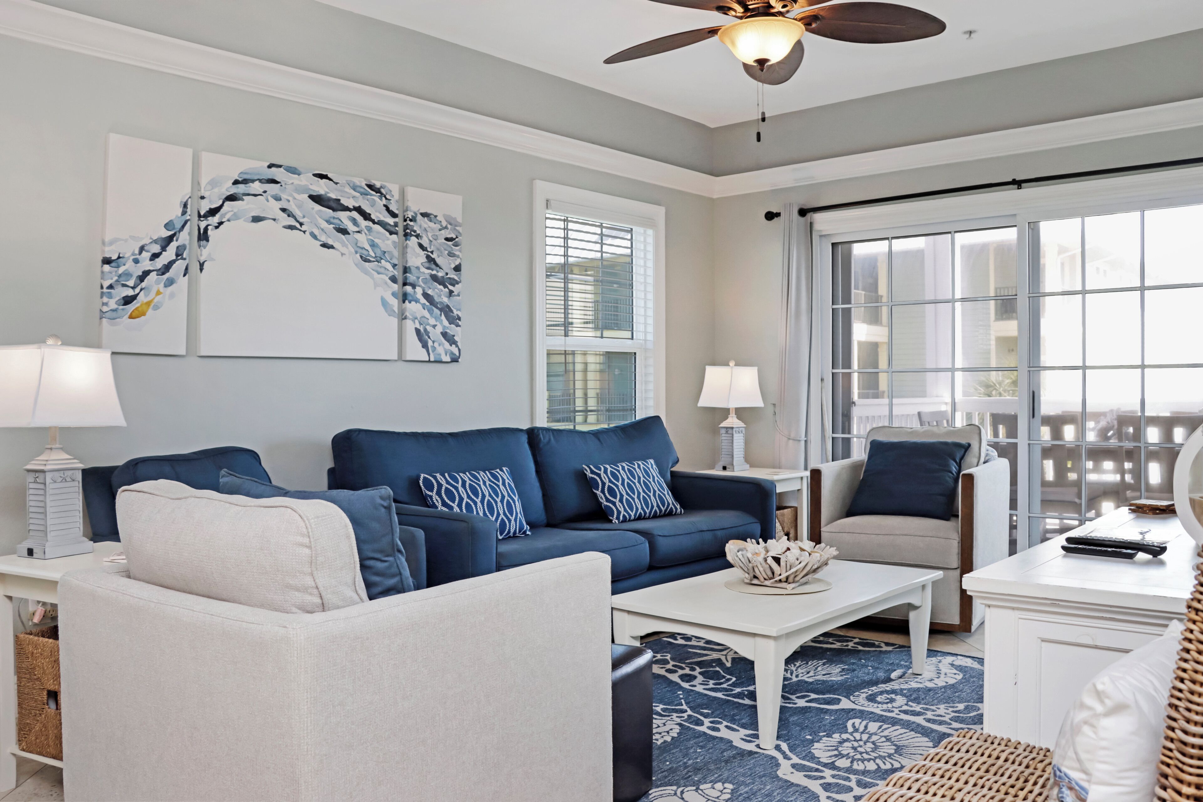 Ein Wohnzimmer mit einem blauen Sofa, einem weißen Sessel und einem Couchtisch. Zwei Wandleuchten, ein Deckenventilator und ein großes Fenster mit Jalousien sind vorhanden.