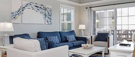 Ein Wohnzimmer mit einem blauen Sofa, einem weißen Sessel und einem Couchtisch. Zwei Wandleuchten, ein Deckenventilator und ein großes Fenster mit Jalousien sind vorhanden.