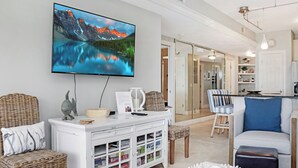 Een woonkamer met een flatscreen televisie, rieten meubels, een witte kast met opbergruimte, en een keuken in de achtergrond.