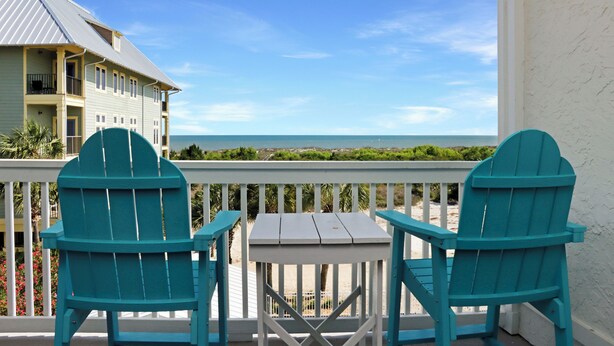 Een balkon met twee turquoise Adirondack-stoelen en een klein tafeltje, met uitzicht op een strand en een gebouw.
