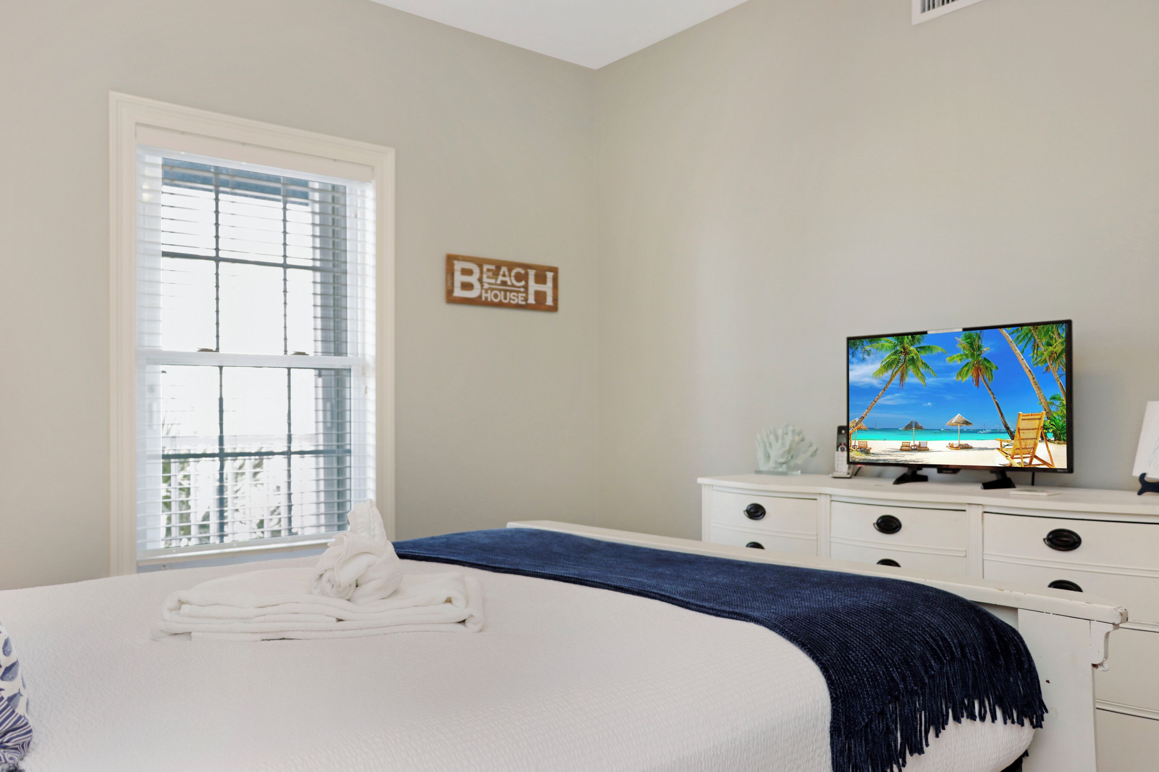 A bedroom with a bed, a TV displaying a beach scene, a window with blinds, and a wall sign that says 'Beach House'.