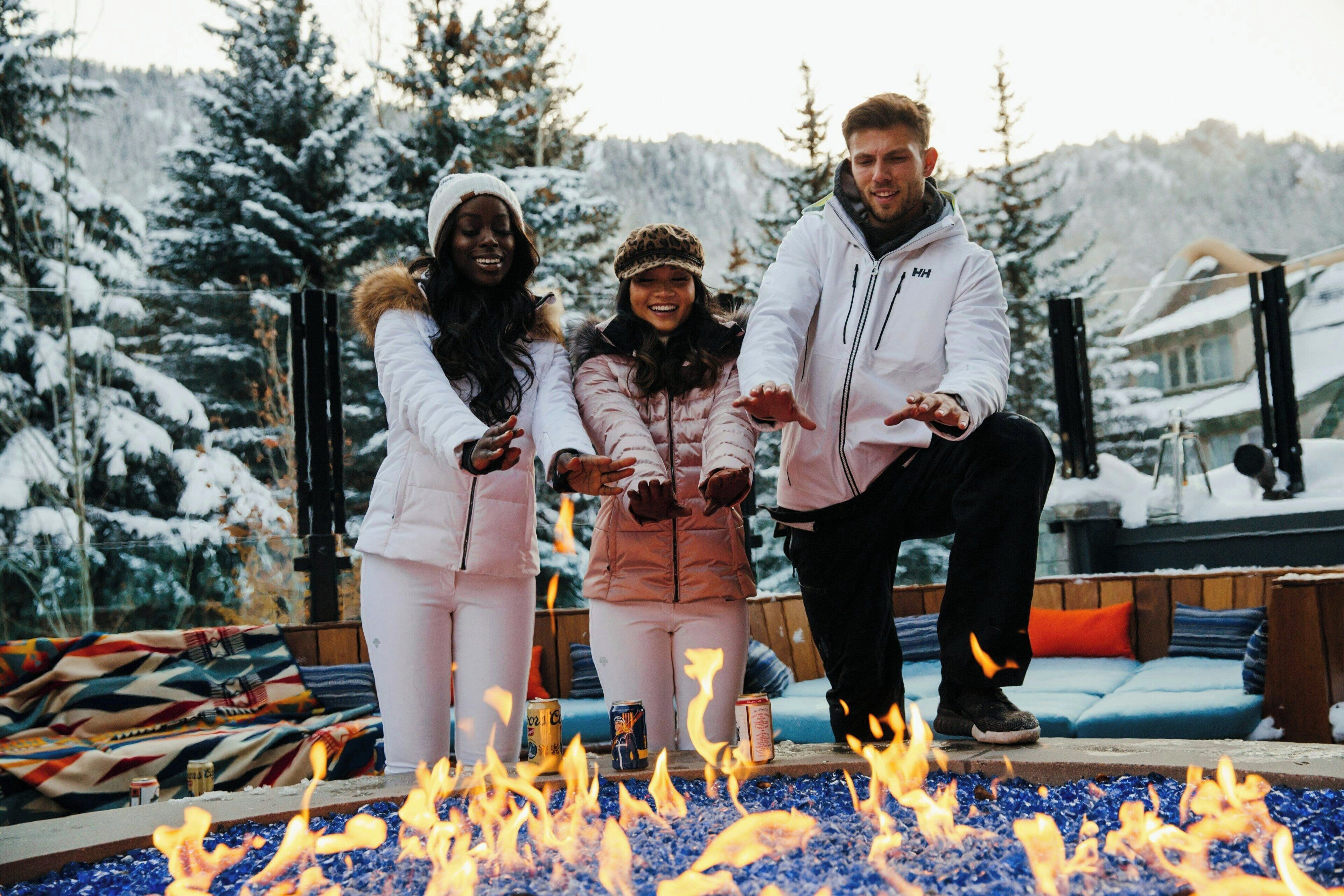 Tres personas junto a una fogata, con árboles cubiertos de nieve al fondo.