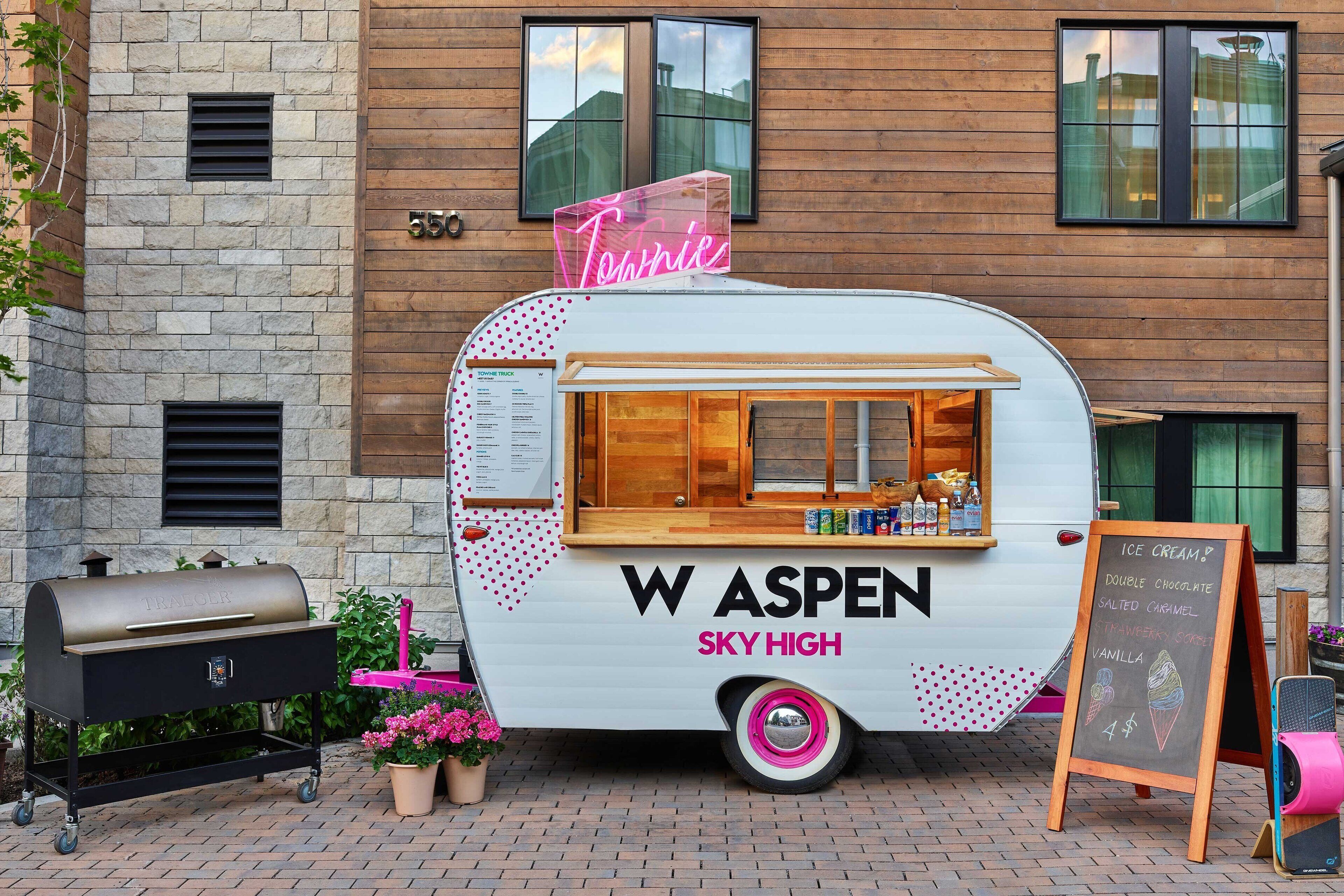 A food truck named 'W ASPEN SKY HIGH' with a neon sign, parked in front of a building with a stone facade and a window.