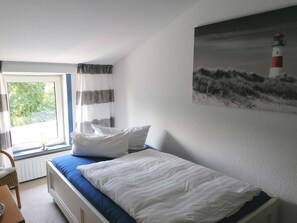 A bedroom with a bed, a window with curtains, a lamp, and a painting of a lighthouse.