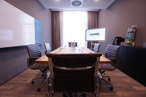A conference room with a large table, four chairs, a screen displaying "HARBR.", and a coffee machine. - Comfort - HARBR. Hotel Heilbronn (Heilbronn)
