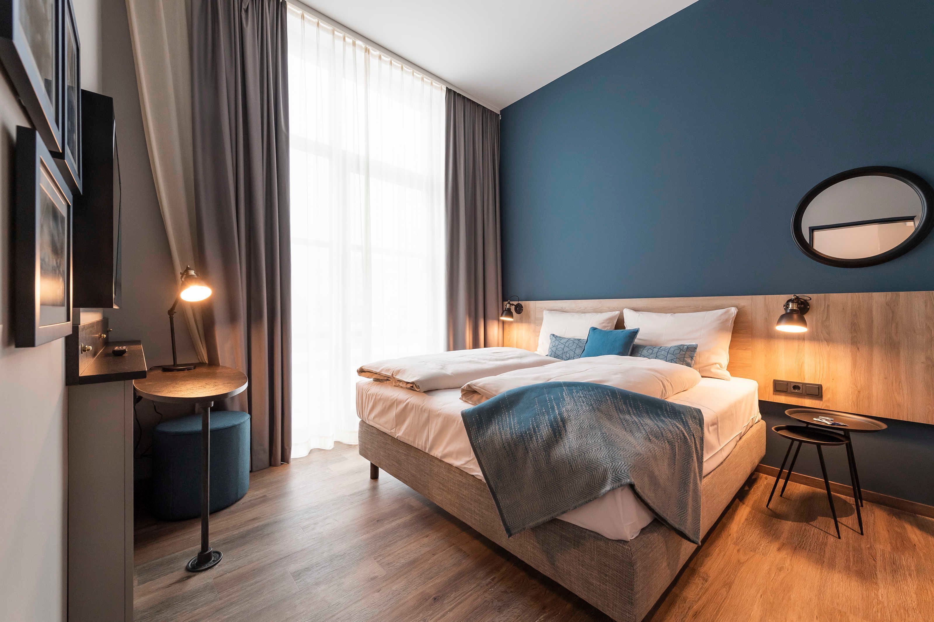 A modern hotel room with a bed, bedside tables, a mirror, and a view of the outdoors.