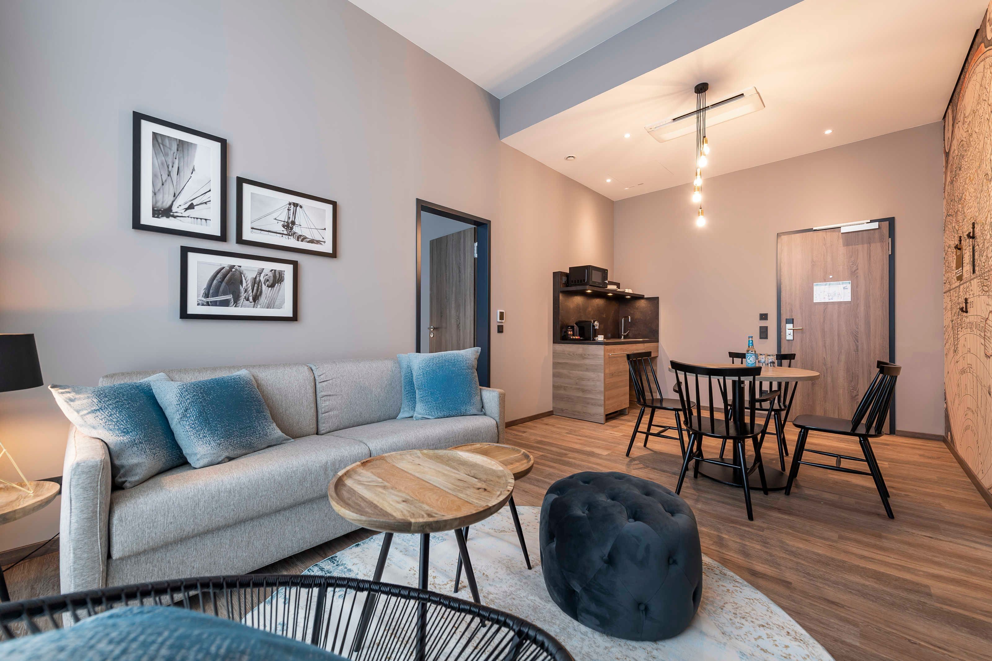 Una sala moderna con sofá, mesa de centro, otomán y comedor con sillas, además de una pequeña cocina.