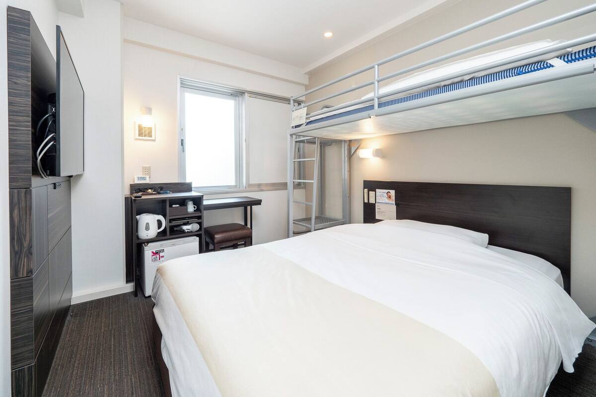 A hotel room with a bunk bed, a desk with a chair, a coffee maker, a clock, and a window with blinds.