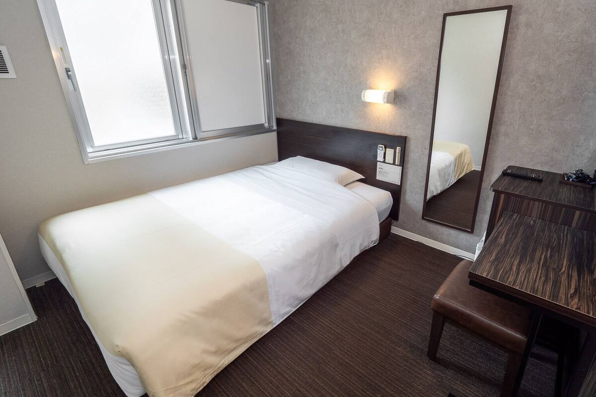 A hotel room with a bed, a bedside table, a mirror, and a window.