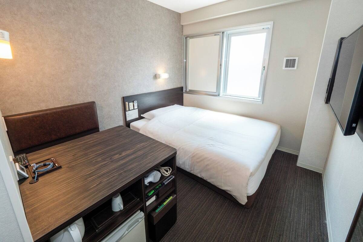 A hotel room with a bed, a desk with a chair, a TV, and a window.