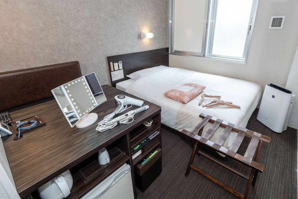 A compact hotel room with a bed, bedside table, mirror, and a small desk with a hairdryer and other items.