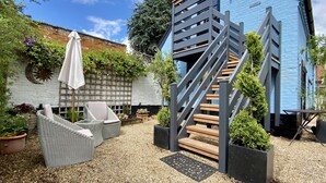 A modern outdoor area with a blue building, a wooden staircase, wicker furniture, and a white umbrella. - A Spacious, Modern & Contemporary Loft Space (Wickham Market)