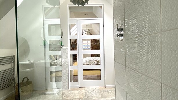 A bathroom with a glass shower enclosure, a white tiled wall, and a white door leading to a bedroom with a bed and a window. - A Spacious, Modern & Contemporary Loft Space (Wickham Market)