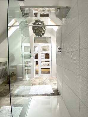 A bathroom with a glass shower enclosure, a white tiled wall, and a white door leading to a bedroom with a bed and a window.