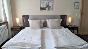 A double bed with two pillows, a nightstand with a lamp, a radiator, and a painting on the wall. - Small double room - Altstadthotel Goldene Kugel Object ID 124185 (Waren (Müritz))