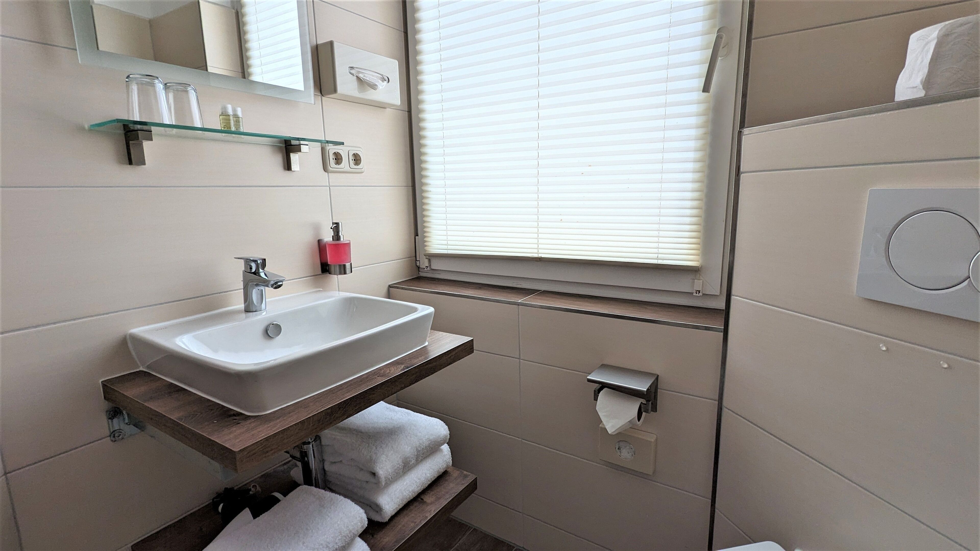 Ein Badezimmer mit einem weißen Waschbecken, einer Glasablage darüber, einem Handtuchhalter, einem Toilettenpapierhalter und einem Fenster mit Jalousien.
