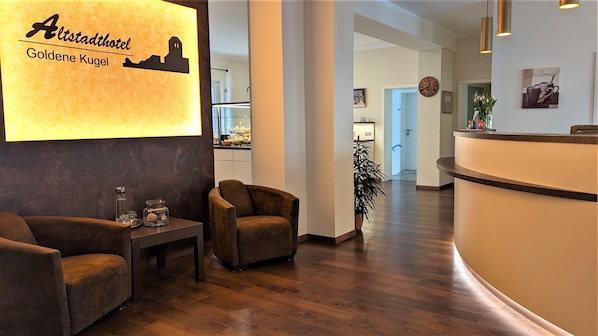 A hotel reception area with a modern design, featuring a reception desk, two armchairs, a small table with decorative items, and a wall-mounted sign with the hotel name and location.