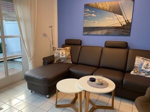 A living room with a sectional sofa, two round tables, a large window with curtains, and a sailboat painting on the wall.