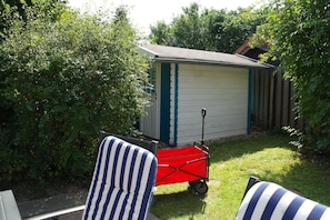 Een klein tuinhuis met een schuin dak, een rode kruiwagen en gestreept tuinmeubilair.