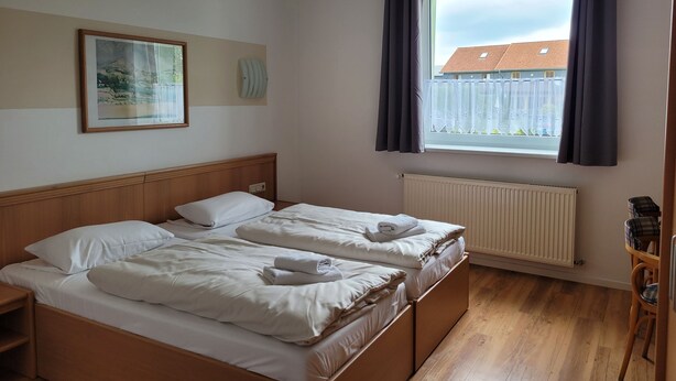 Ein Schlafzimmer mit zwei Betten, einem Fenster mit Vorhängen, einem Heizkörper, einem Holzboden und einem gerahmten Bild an der Wand.