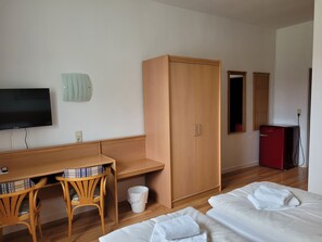 Una habitación con un escritorio de madera, dos sillas, un televisor, un armario, una cama con toallas, un cubo de basura y un refrigerador rojo.