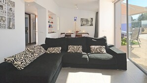 A modern living room with a black sectional sofa, a dining area with a table and chairs, and a large sliding glass door leading to an outdoor patio. - DJ's House - Modern and stylish villa by the sea in Mallorca (Son Serra de Marina)