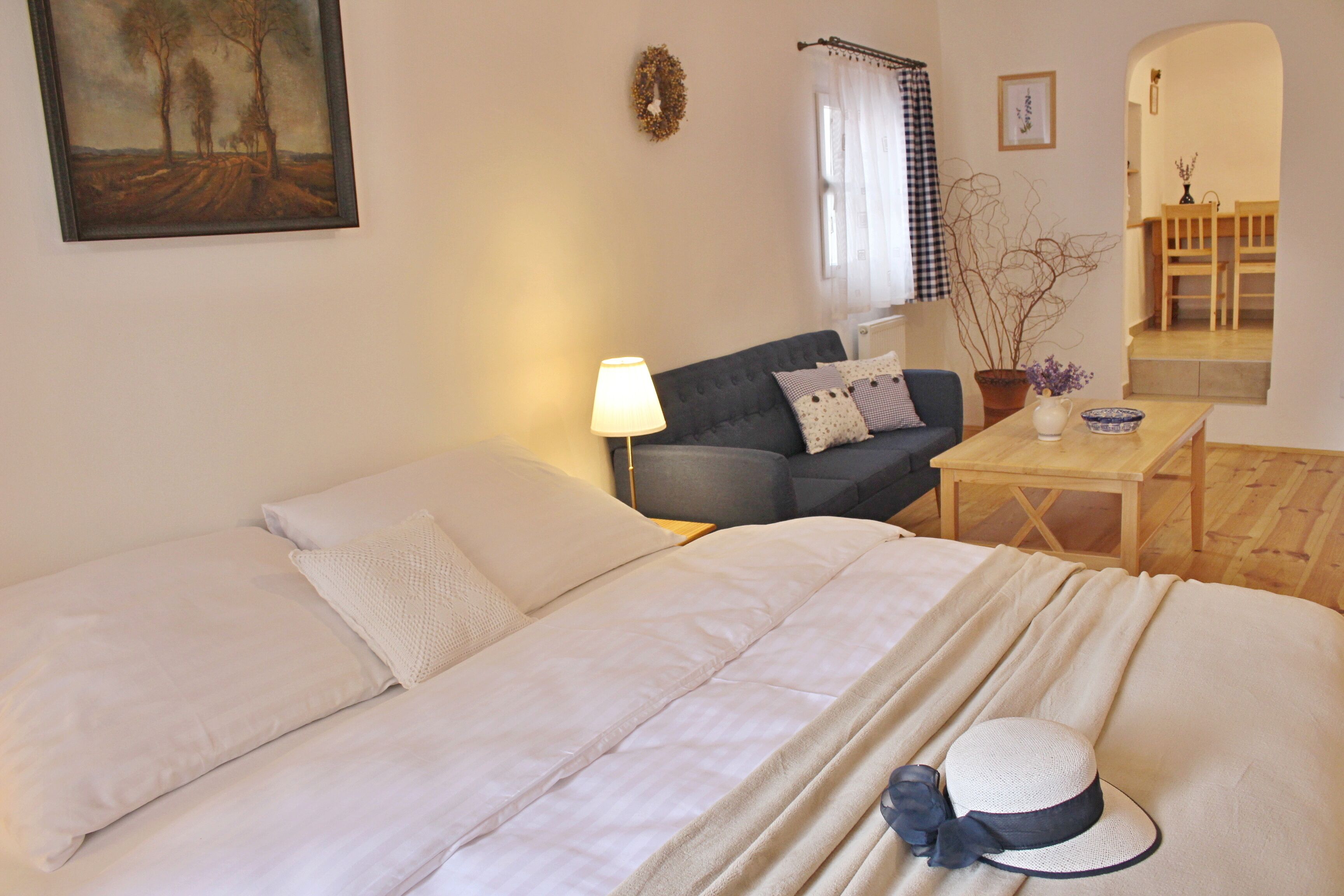 A neatly arranged bedroom with a bed, a sofa, a small table with a lamp, and a decorative hat on the bed.