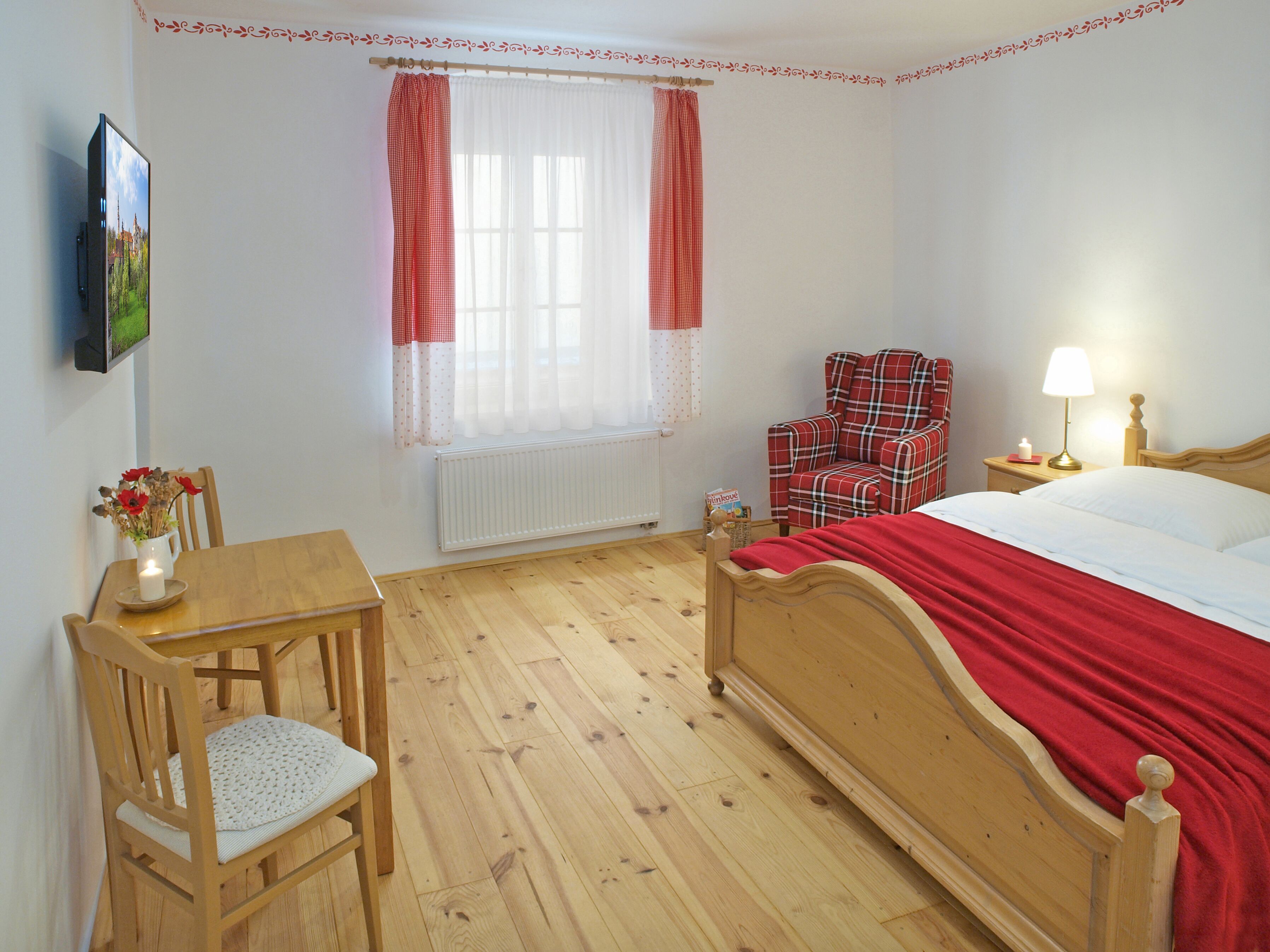 Ein Schlafzimmer mit einem hölzernen Bett, einem Sessel, einem Tisch, einem Fenster mit Vorhängen, einem Heizkörper und einem an der Wand befestigten Fernseher.