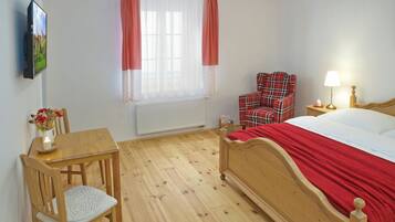 Ein Schlafzimmer mit einem hölzernen Bett, einem Sessel, einem Tisch, einem Fenster mit Vorhängen, einem Heizkörper und einem an der Wand befestigten Fernseher.