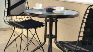 Un balcón con dos sillas de alambre negras y una mesita redonda. Sobre la mesa hay una taza y un plato, y un libro titulado “HIGH TREK” a su lado. El fondo muestra un paisaje urbano con montañas.