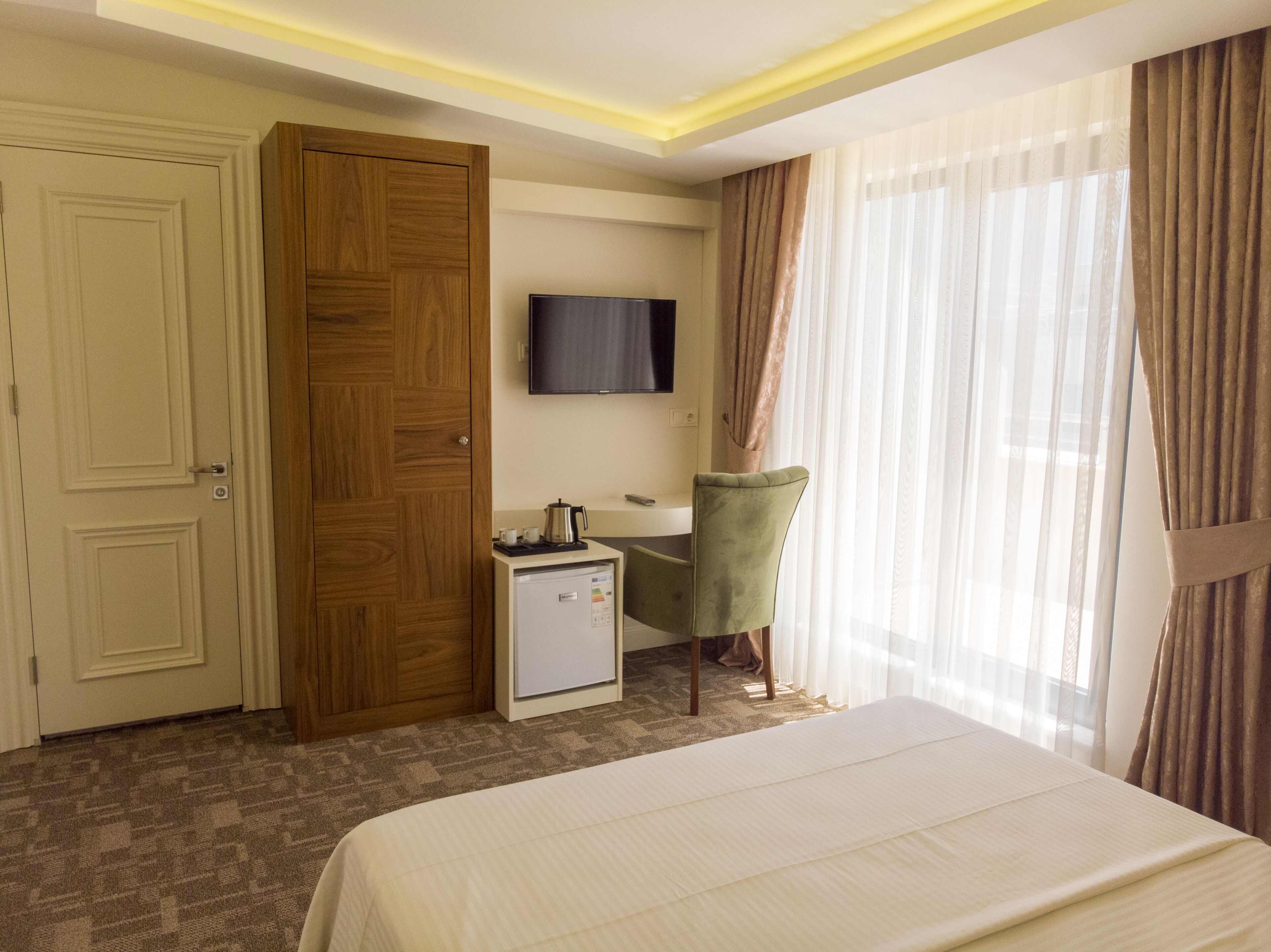 A hotel room with a bed, a desk with a television, a minibar, and a green chair.