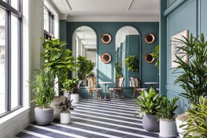 Un couloir orné de plantes en pot, doté d’un sol à carreaux et offrant une vue sur une pièce où se trouvent un grand miroir et un tableau.