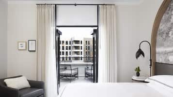 Una camera d'hotel con un letto, una poltrona scura, un tavolino con una pianta e vista su un edificio da una grande finestra.