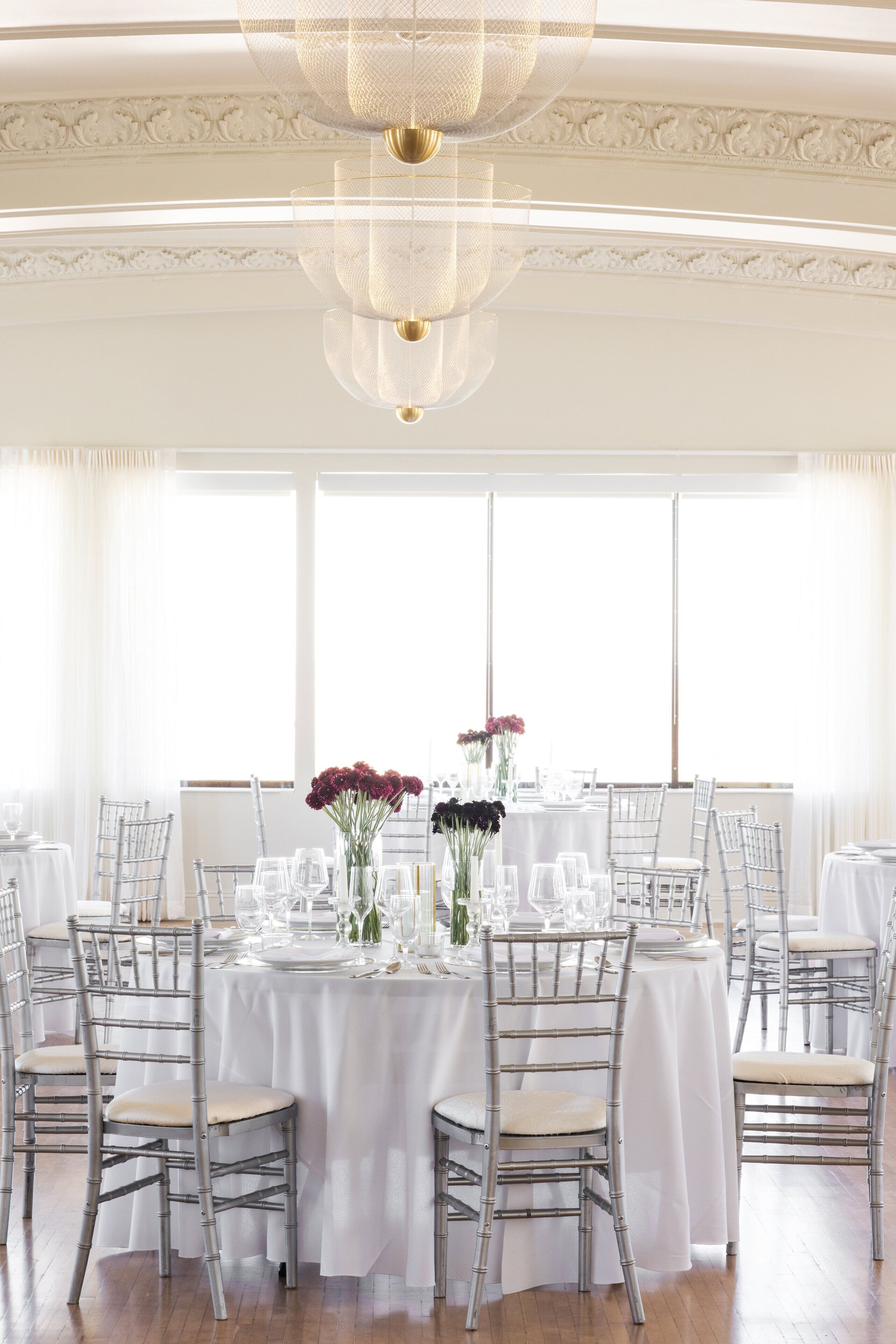 A dining area with white tablecloths, clear chairs, and a chandelier.