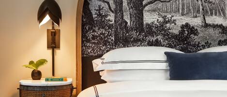 Una camera da letto con un letto, una lampada a parete, un tavolino rotondo con una pianta e un grande arazzo raffigurante una scena di foresta.