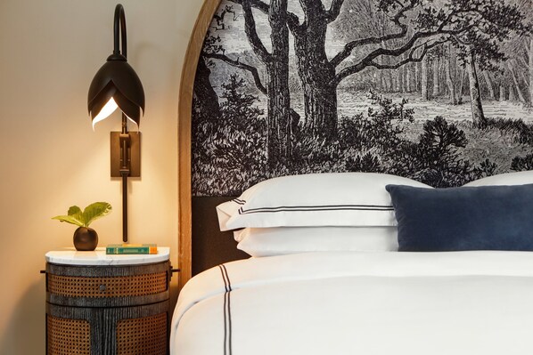 Un dormitorio con una cama, una lámpara de pared, una pequeña mesa redonda con una planta y un tapiz grande que representa un paisaje boscoso.