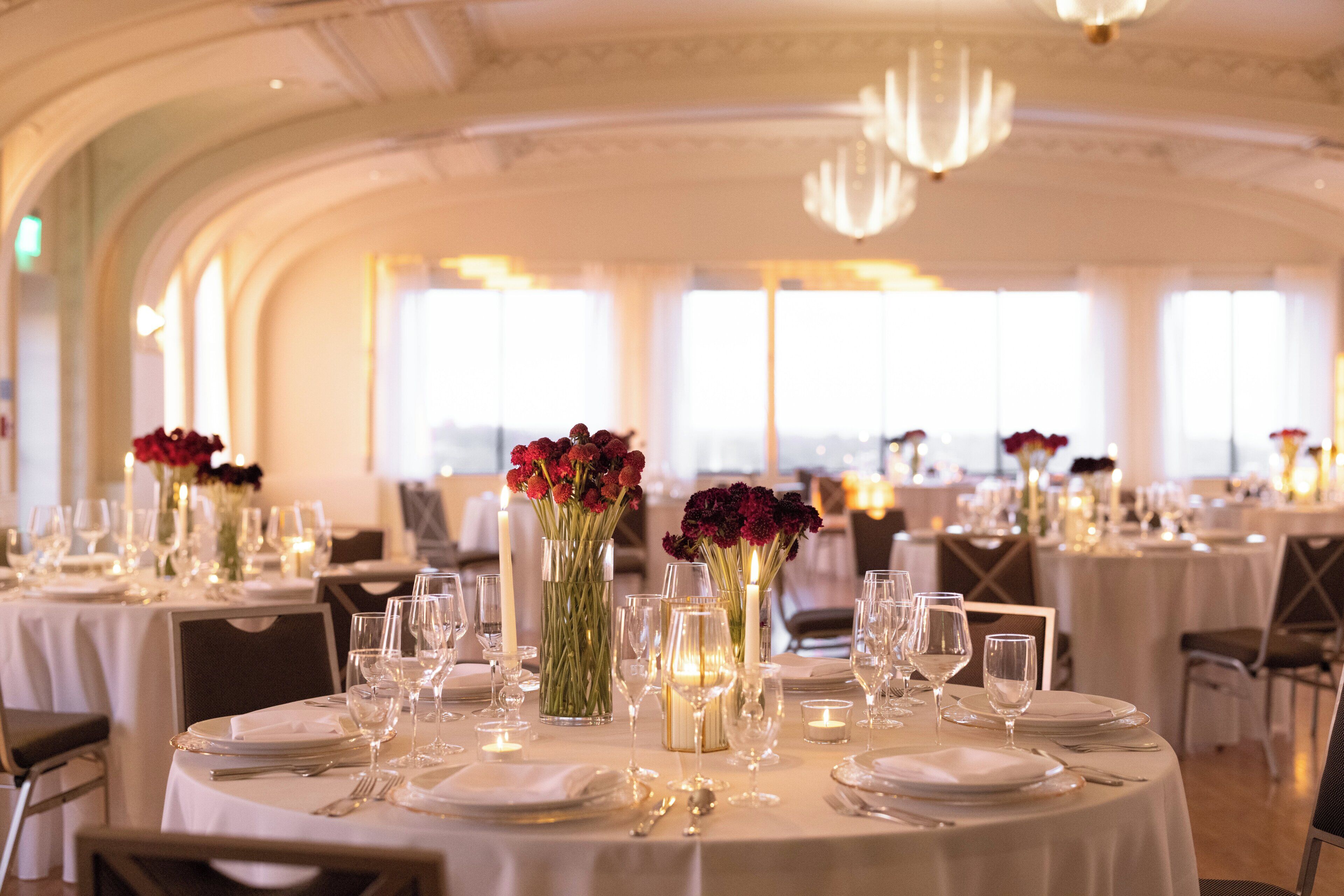 A banquet hall with round tables set for dining, featuring glassware, plates, and candles.