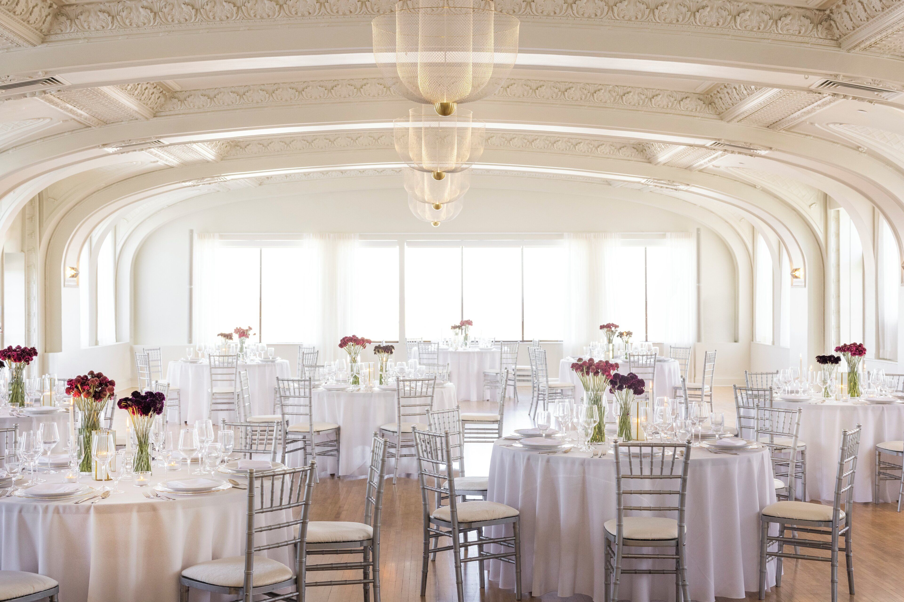 A banquet hall with multiple round tables, each set with white tablecloths, chairs, and floral centerpieces.
