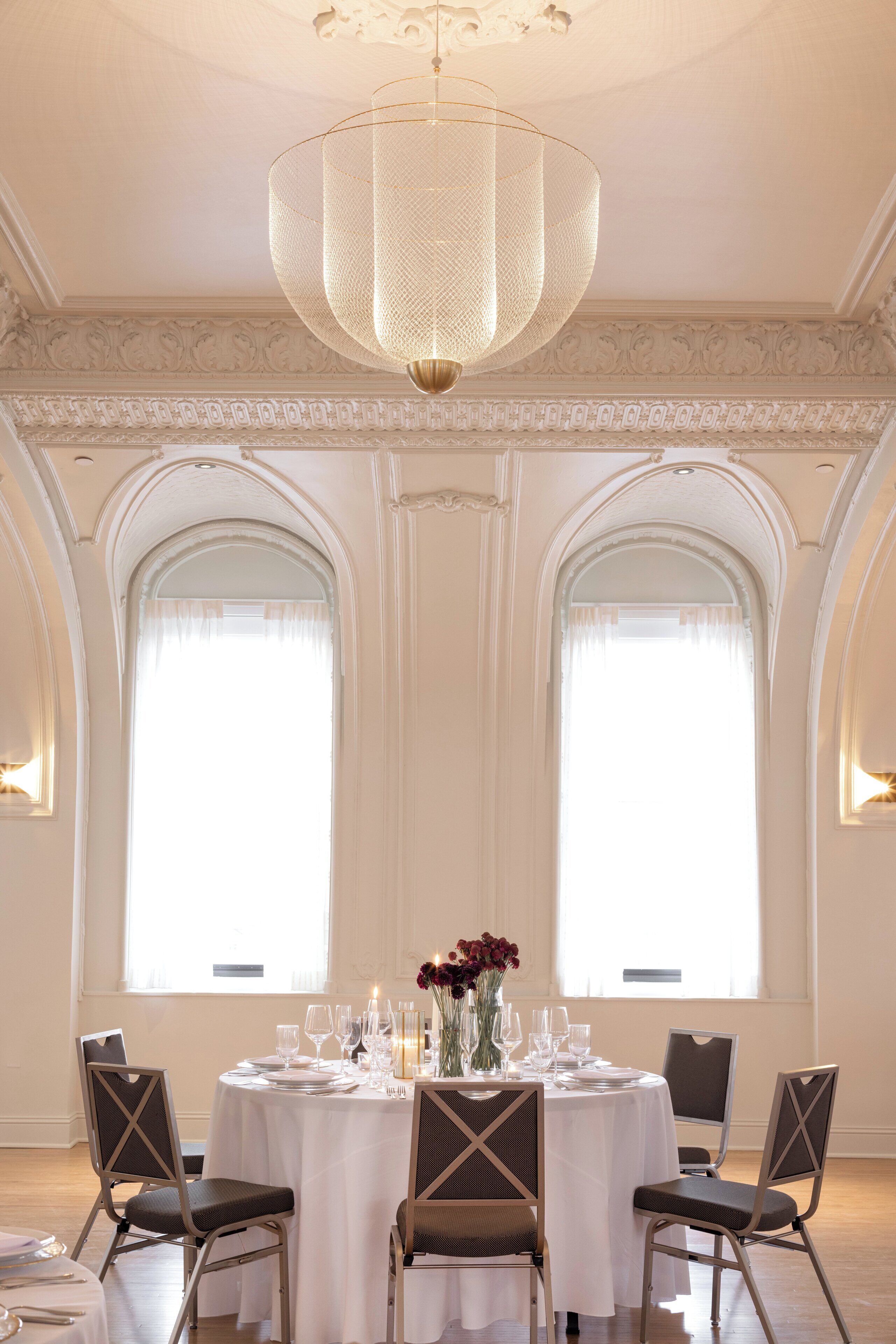 A dining room with a round table set for six, flanked by two large windows with curtains, a chandelier, and ornate wall moldings.