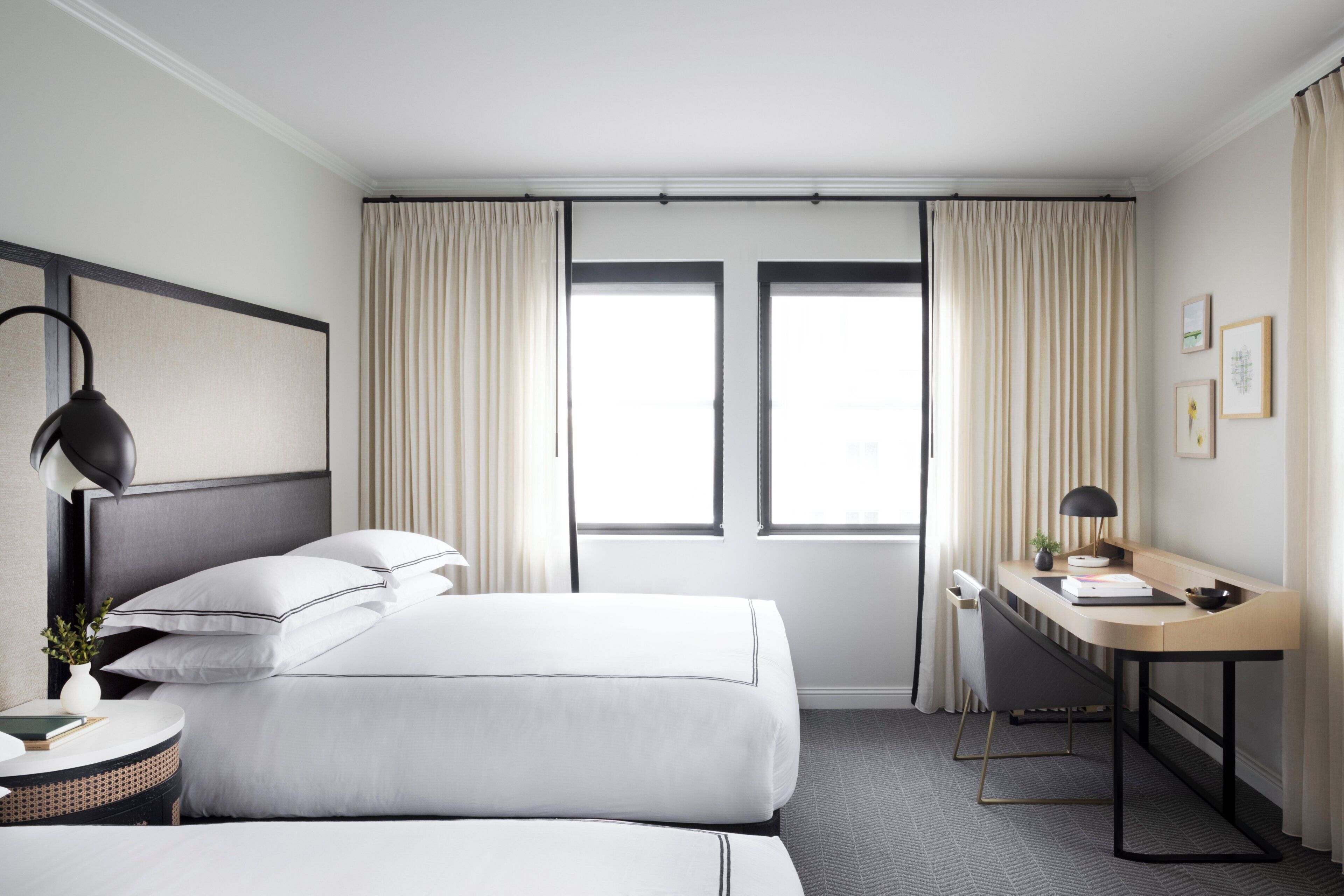 Habitación de hotel con dos camas, un escritorio, una silla, una lámpara y un ventanal con cortinas.