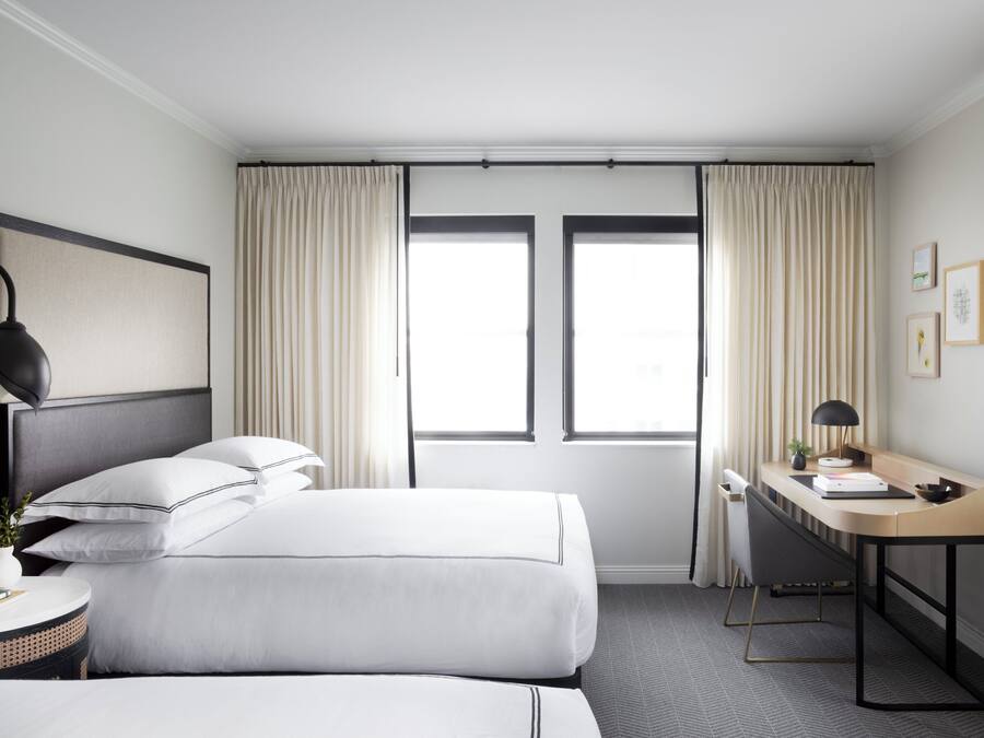 Habitación de hotel con dos camas, un escritorio, una silla, una lámpara y un ventanal con cortinas.