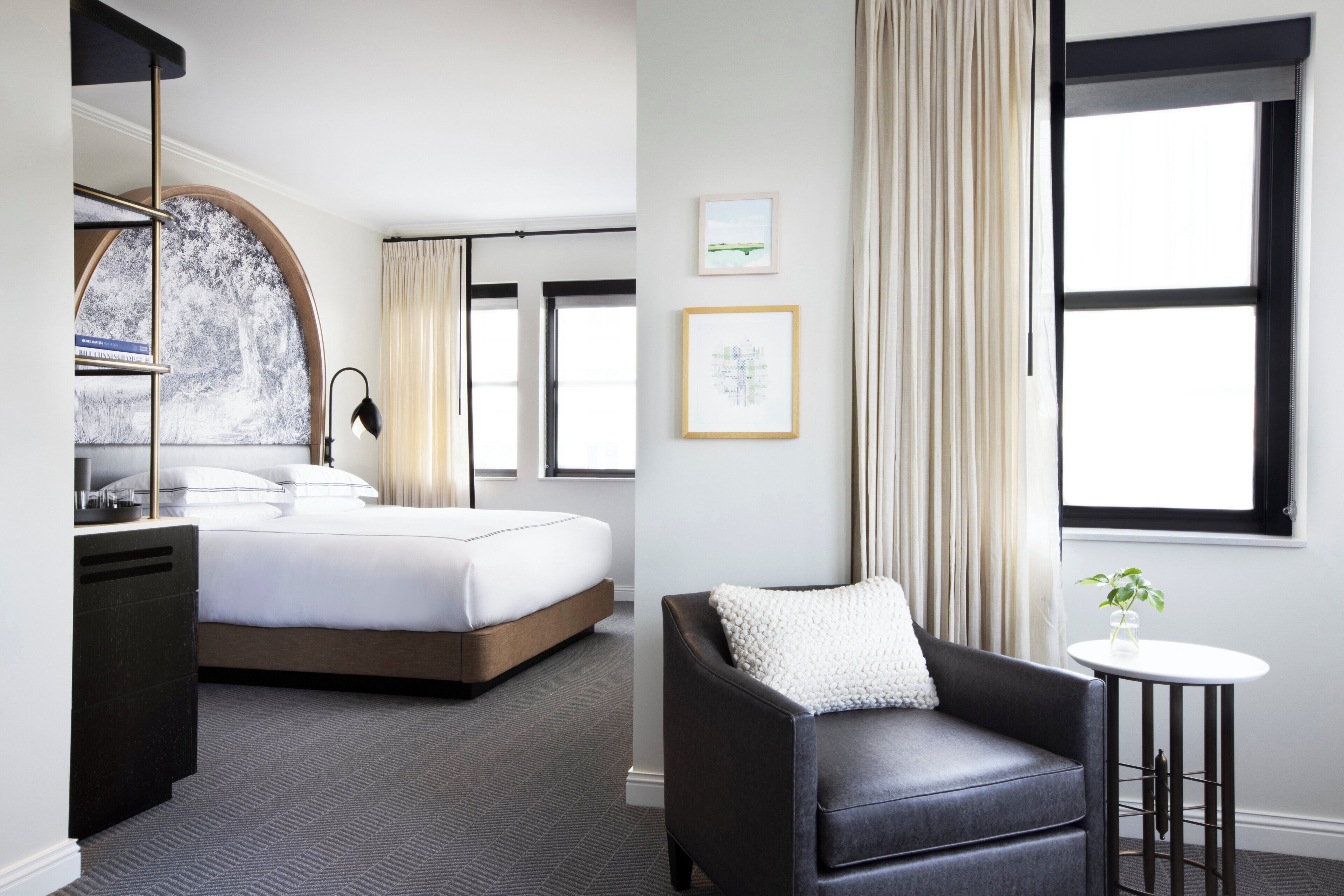 Ein Hotelzimmer mit einem Bett, einem Schreibtisch, einem Sessel, einem kleinen Tisch mit Pflanze und zwei mit Vorhängen versehenen Fenstern.