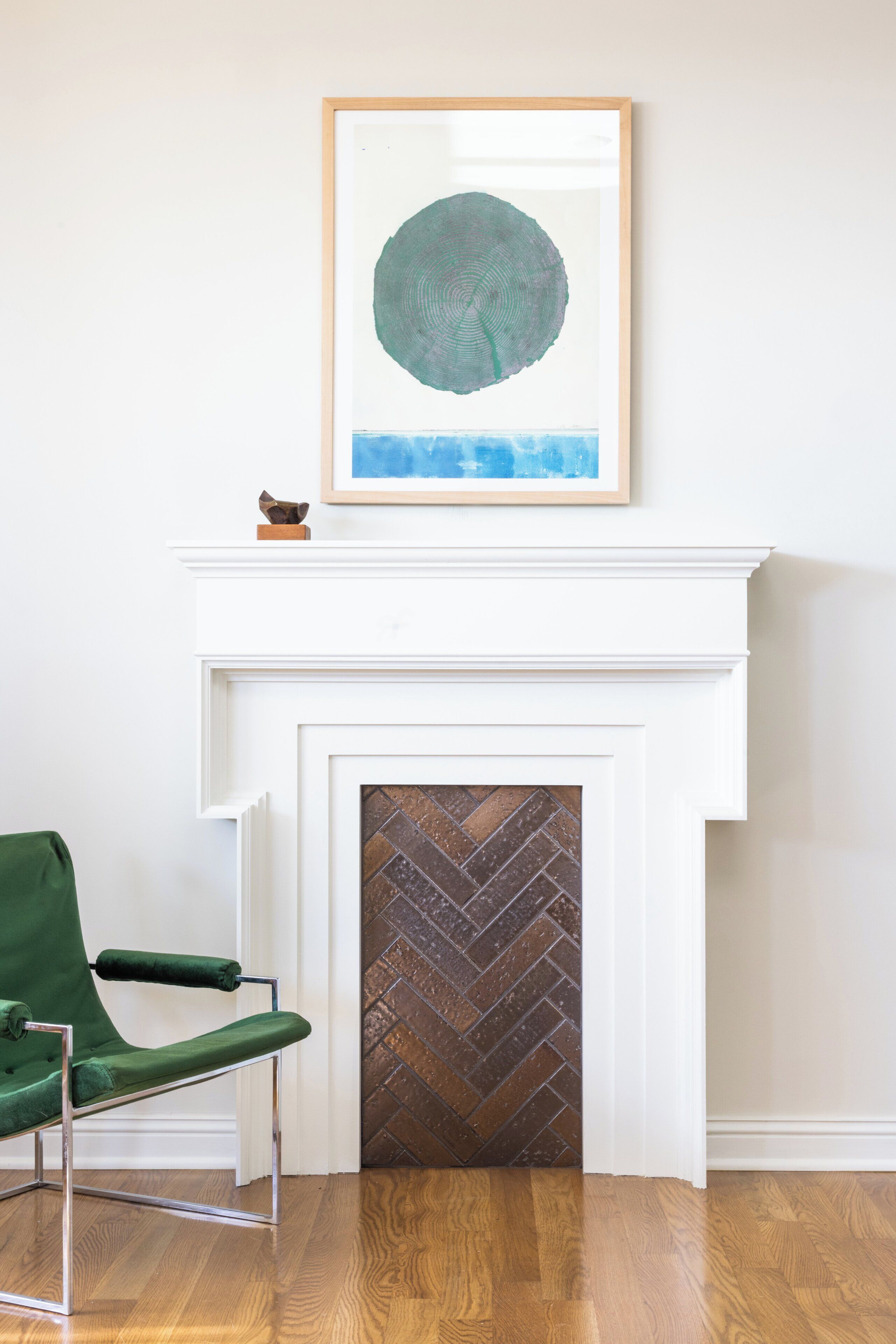 A room with a green chair, a fireplace with a herringbone-patterned door, and a framed artwork above the mantel.