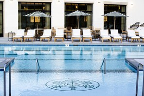 Une piscine entourée de chaises longues et de tables, le tout adossé à un bâtiment doté de grandes fenêtres.